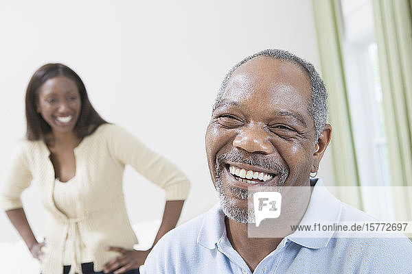 Porträt eines lächelnden Mannes mittleren Alters mit einer im Hintergrund stehenden Frau mittleren Alters mit ausgebreiteten Armen
