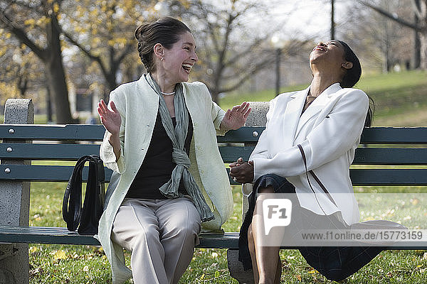 Zwei Frauen sitzen auf einer Bank im Park und lachen.