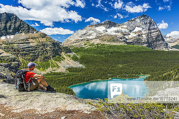Male hiker sitting on rocky ridge overlooking blue alpine lake and mountains in the distance with blue sky and clouds  Yoho National Park; Field  British Columbia  Canada