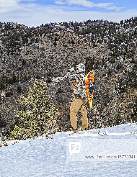 Hunter with snowshoes and rifle standing on a hillside and looking out with binoculars; Denver  Colorado  United States of America