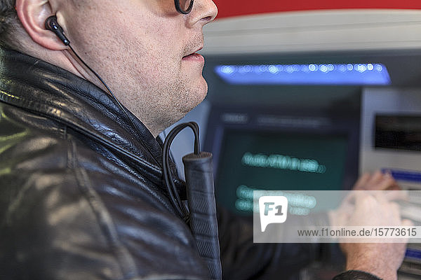 Mann mit Sehbehinderung  der mit einem weißen Stock und Kopfhörern an einem Bankautomaten steht