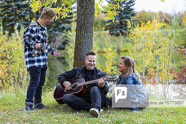 A young man with Down Syndrome playing a tambourine while his father plays a guitar and his mother sings along while enjoying each other's company in a city park on a warm fall evening: Edmonton  Alberta  Canada