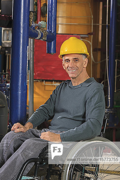 Porträt eines Industriearbeiters im Rollstuhl  der einen gelben Schutzhelm trägt und in die Kamera lächelt