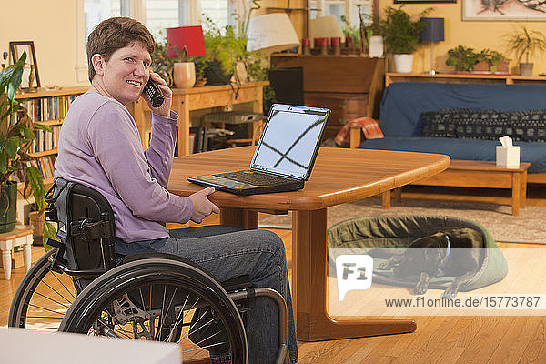 Behinderte Person im Rollstuhl  die zu Hause einen Laptop benutzt und mit ihrem Hund im Hintergrund telefoniert