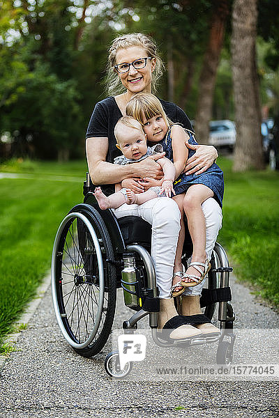 Eine querschnittsgelähmte Mutter  die ihre Töchter auf dem Schoß trägt  während sie einen Rollstuhl benutzt  posiert an einem warmen Sommernachmittag im Freien für die Kamera: Edmonton  Alberta  Kanada