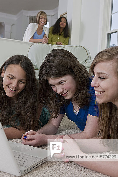 Drei Mädchen im Teenageralter benutzen einen Laptop  während ihre Mütter ihnen über die Schultern schauen