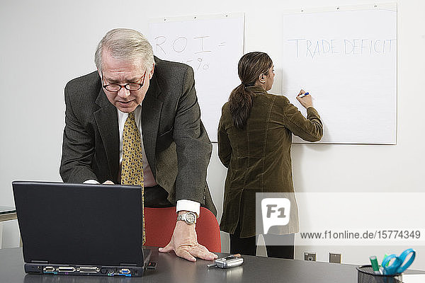 Eine Geschäftsfrau schreibt an einer weißen Tafel  während der CEO am Laptop steht.