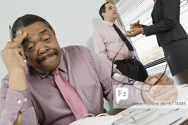 Businessman sitting in an office and looking stressed with his colleagues talking in the background