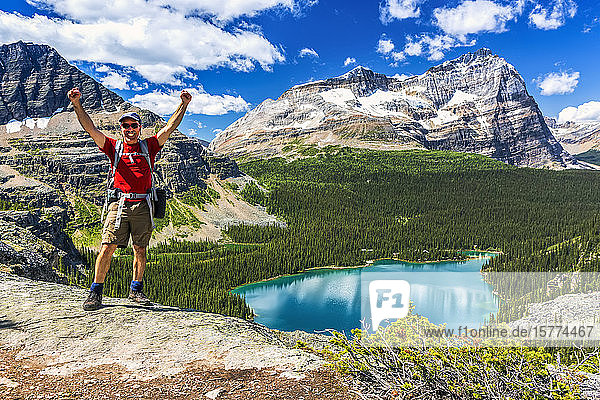 Male hiker standing with arms in the air on rocky ridge overlooking blue alpine lake and mountains in the distance with blue sky and clouds  Yoho National Park; Field  British Columbia  Canada