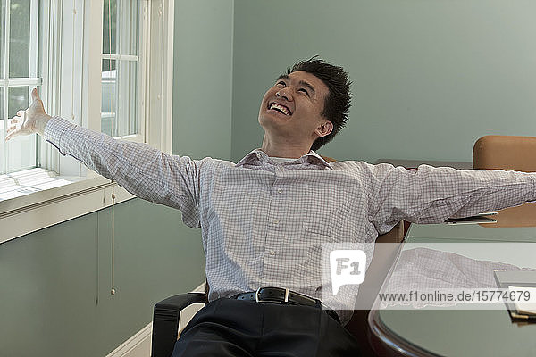 Junger asiatischer Geschäftsmann sitzt auf einem Stuhl am Arbeitsplatz und feiert einen Sieg