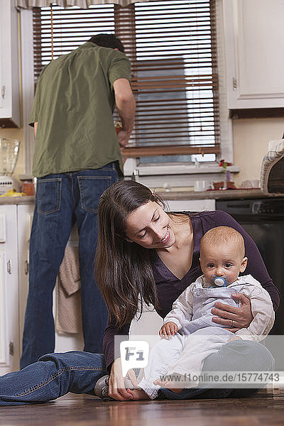 Eine junge Mutter hält ihren kleinen Sohn  während der Vater in der Küche im Hintergrund steht.