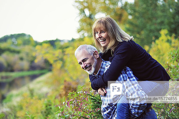 Ein reifer Mann  der sich amüsiert und seine Frau Huckepack auf einem Weg entlang eines Flusses in einem Stadtpark an einem warmen Herbstabend nimmt; St. Albert  Alberta  Kanada