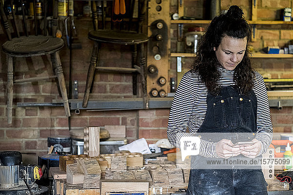 Frau mit langen braunen Haaren  die eine Latzhose trägt und in einer Holzwerkstatt steht und ein Mobiltelefon benutzt.