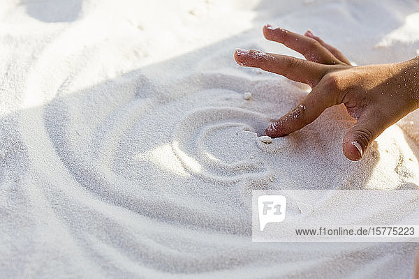 Ein Junge  der mit seinen Händen in den weißen Sand zeichnet.