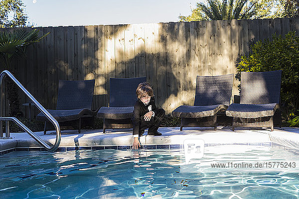 A young boy wearing black suit and bow tie standing by a swimming pool