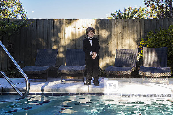 A young boy wearing black suit and bow tie standing by a swimming pool