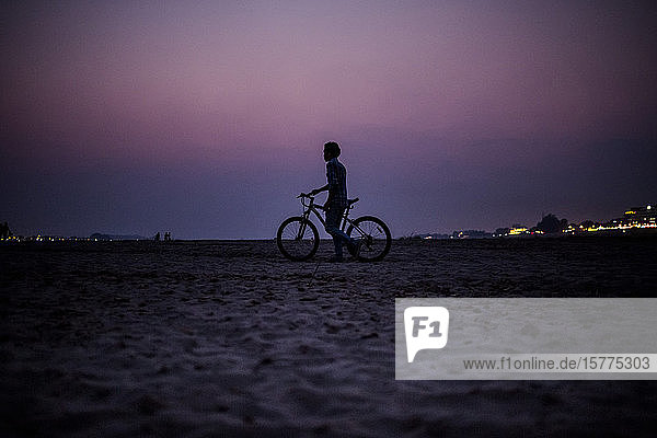 Silhouette eines Jungen  der bei Sonnenuntergang am Ufer eines Flusses ein Fahrrad schiebt.