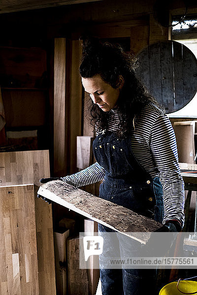 Frau mit langen braunen Haaren in Latzhose in Holzwerkstatt stehend  ein Stück Holz haltend.