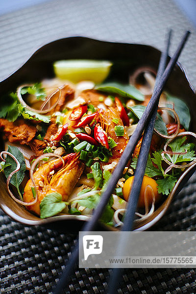 Hochwinkel-Nahaufnahme von Essstäbchen auf einer Schüssel mit asiatischem Essen mit Nudeln  Garnelen  Gemüse und Chili-Garnitur.