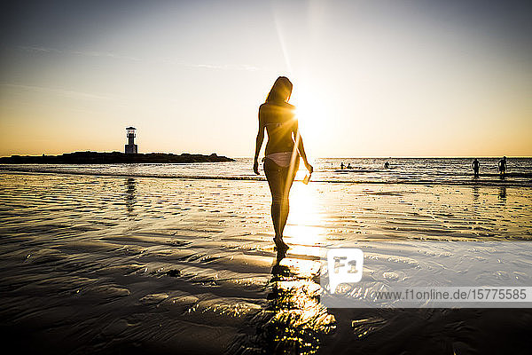 Rückansicht einer Frau  die bei Sonnenuntergang in Richtung Strand geht.