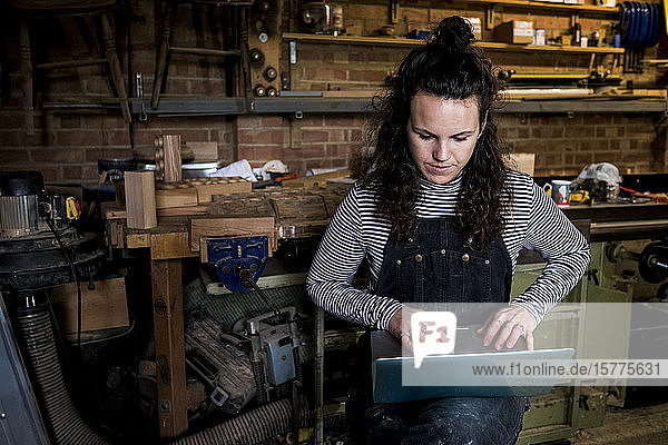 Frau mit langen braunen Haaren in Latzhose  die in einer Holzwerkstatt steht und am Laptop tippt.