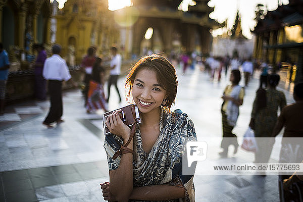 Junge Frau steht auf dem Stadtplatz  hält eine alte Polaroidkamera in der Hand und lächelt in die Kamera.