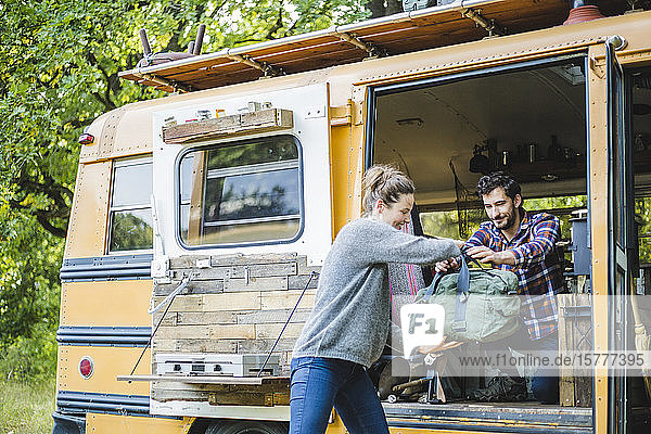 Lächelnde Frau hilft Mann beim Ausladen des Gepäcks aus gelbem Wohnmobil beim Camping im Wald