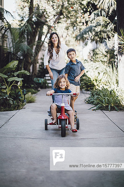 Tochter fährt Dreirad  während Mutter und Bruder im Hintergrund stehen