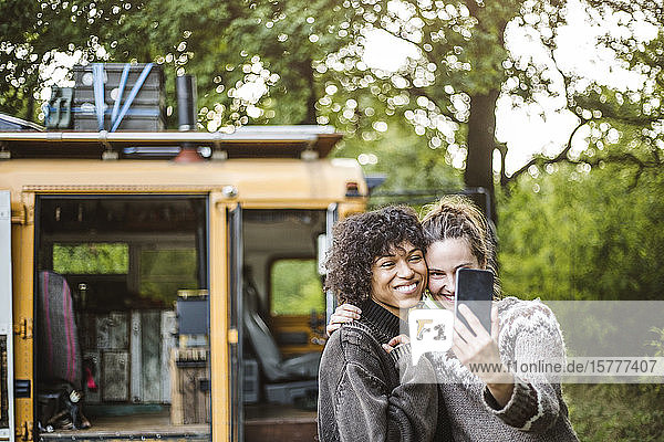 Frau nimmt Selfie mit Freundin gegen Wohnwagen beim Camping im Wald