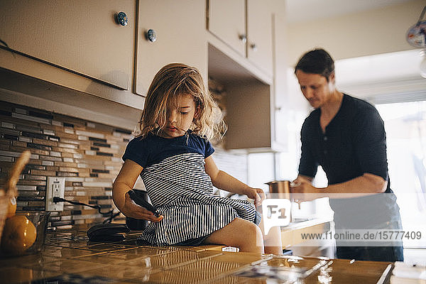 Daughter using phone while father working in kitchen