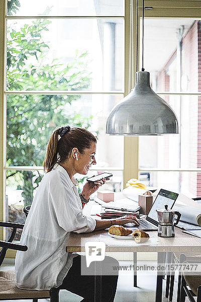 Lächelnde Freiberuflerin benutzt Laptop  während sie im Heimbüro über ein Smartphone spricht