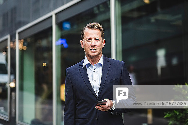 Portrait of mature businessman with smart phone standing against office building