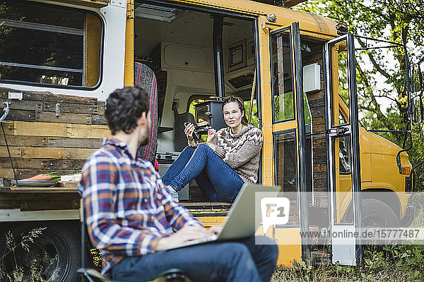 Frau sitzt am Eingang des Wohnwagens  während sie sich beim Zelten im Wald mit einem Freund am Laptop unterhält