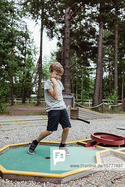 Fröhlicher Junge in voller Länge beim Minigolfspielen im Hinterhof während des Urlaubs