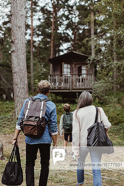 Rückansicht einer Familie mit Taschen  die auf ein Haus im Wald zuläuft