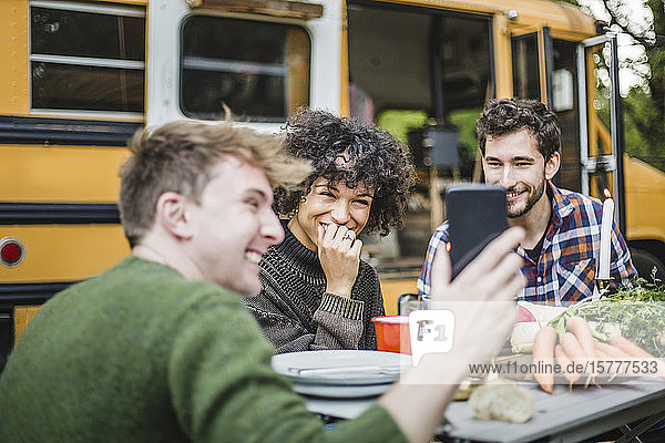 Lächelnder Mann zeigt Freunden  die am Tisch gegen die Karawane sitzen  sein Smartphone