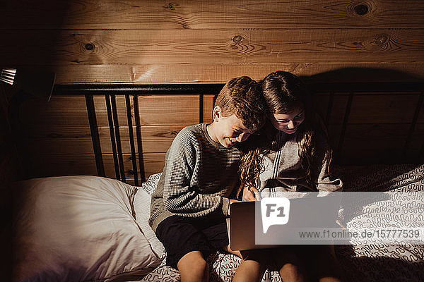 Lächelnde Geschwister mit Laptop  während sie zu Hause im Bett an der Wand sitzen
