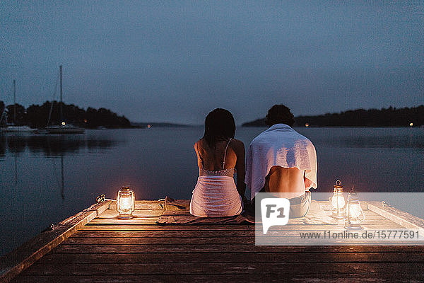 Rear view of couple sitting around illuminated lantern on pier by sea against sky