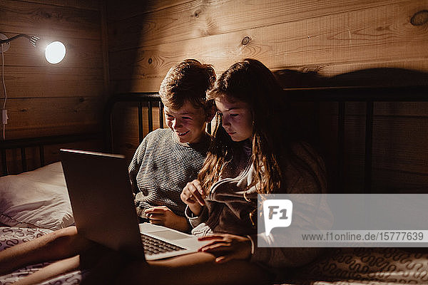 Smiling sibling using laptop while sitting on bed against wall at home