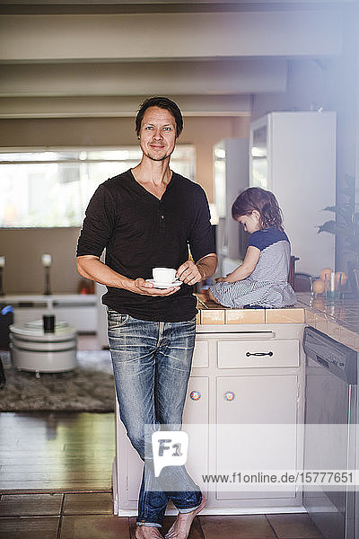 Porträt eines reifen Mannes mit Tasse  während die Tochter auf dem Küchentisch sitzt
