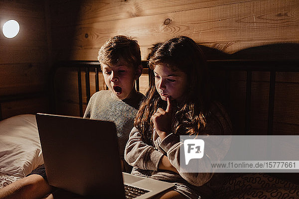 Sibling using laptop while sitting on bed against wall in bedroom