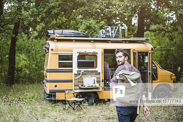 Rückansicht Porträt eines jungen Mannes  der während des Campens mit Rucksack in Richtung Wohnmobil im Wald läuft
