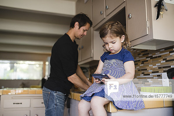 Daughter using phone while father working in domestic kitchen