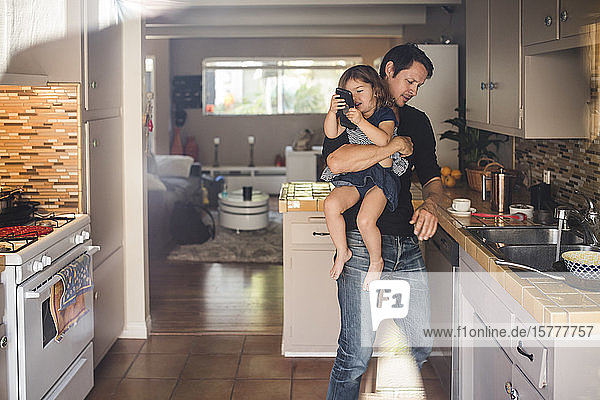 Daughter using phone while father carrying and working in kitchen