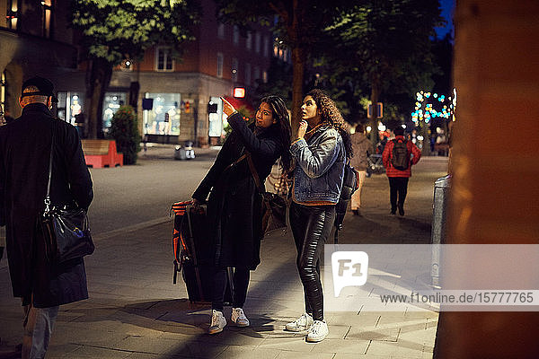 Lesbisches Paar in voller Länge mit Gepäck steht nachts in der Stadt auf dem Bürgersteig