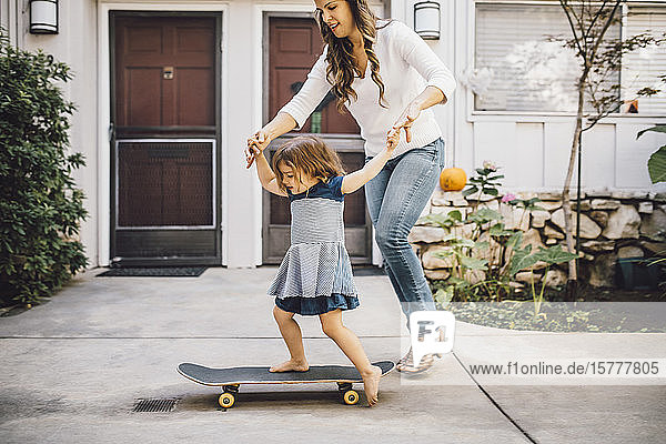 Smiling daughter balancing over skateboard with help of mother on footpath