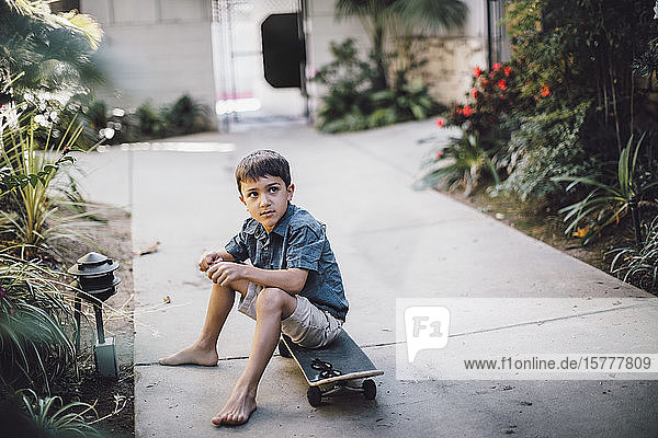 Boy looking away while sitting over skateboard on footpath