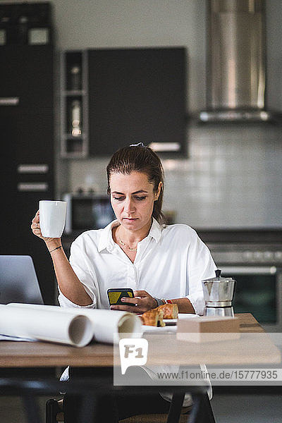 Weibliche Freiberuflerin telefoniert  während sie mit Kaffeetasse zu Hause sitzt