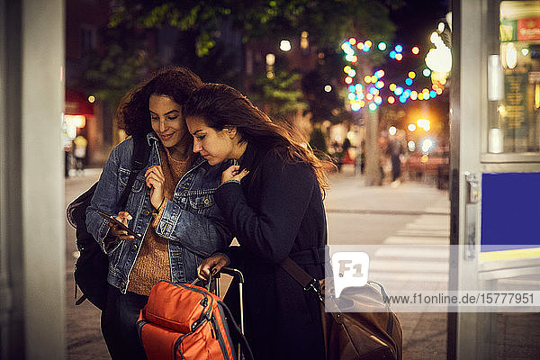 Lesbisches Paar mit Gepäck telefoniert nachts in der Stadt mit dem Handy