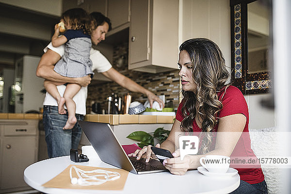 Mother using laptop while father carrying daughter and working in kitchen